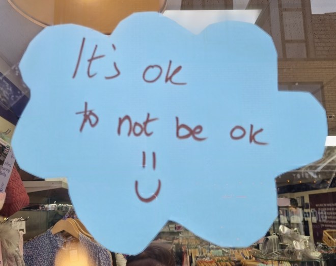 A blue cut out cloud with the words 'It's OK not to be OK' and a smiley face stuck in the Mind Cambridge window for Time To Talk Day 2025